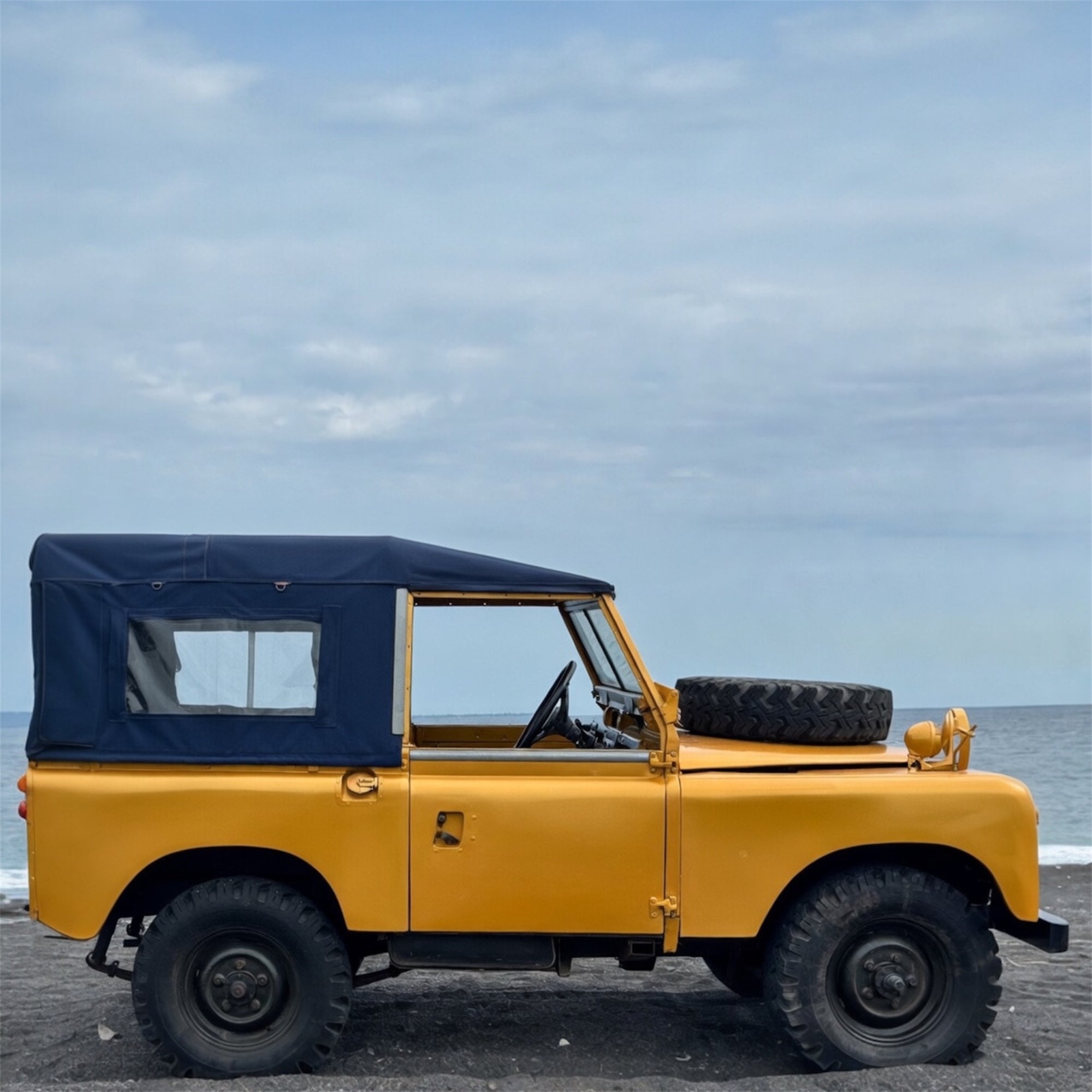 Yellow vintage jeep with a blue roof on a beach with ocean and sky in the background