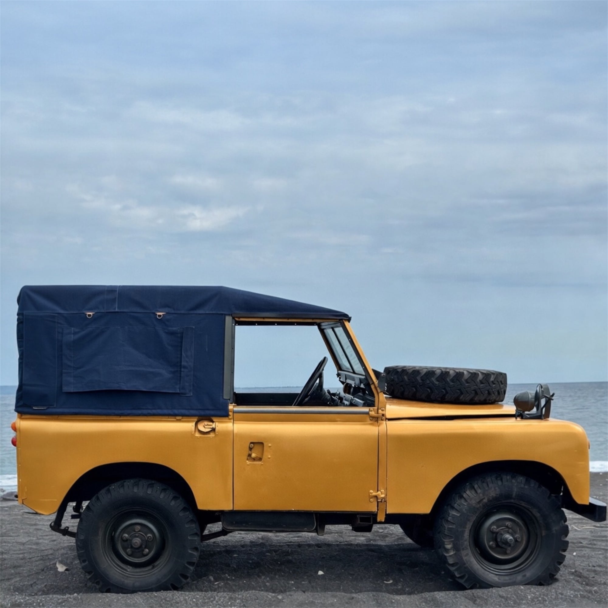 Yellow vintage jeep with a blue soft top on a beach with ocean and sky in the background